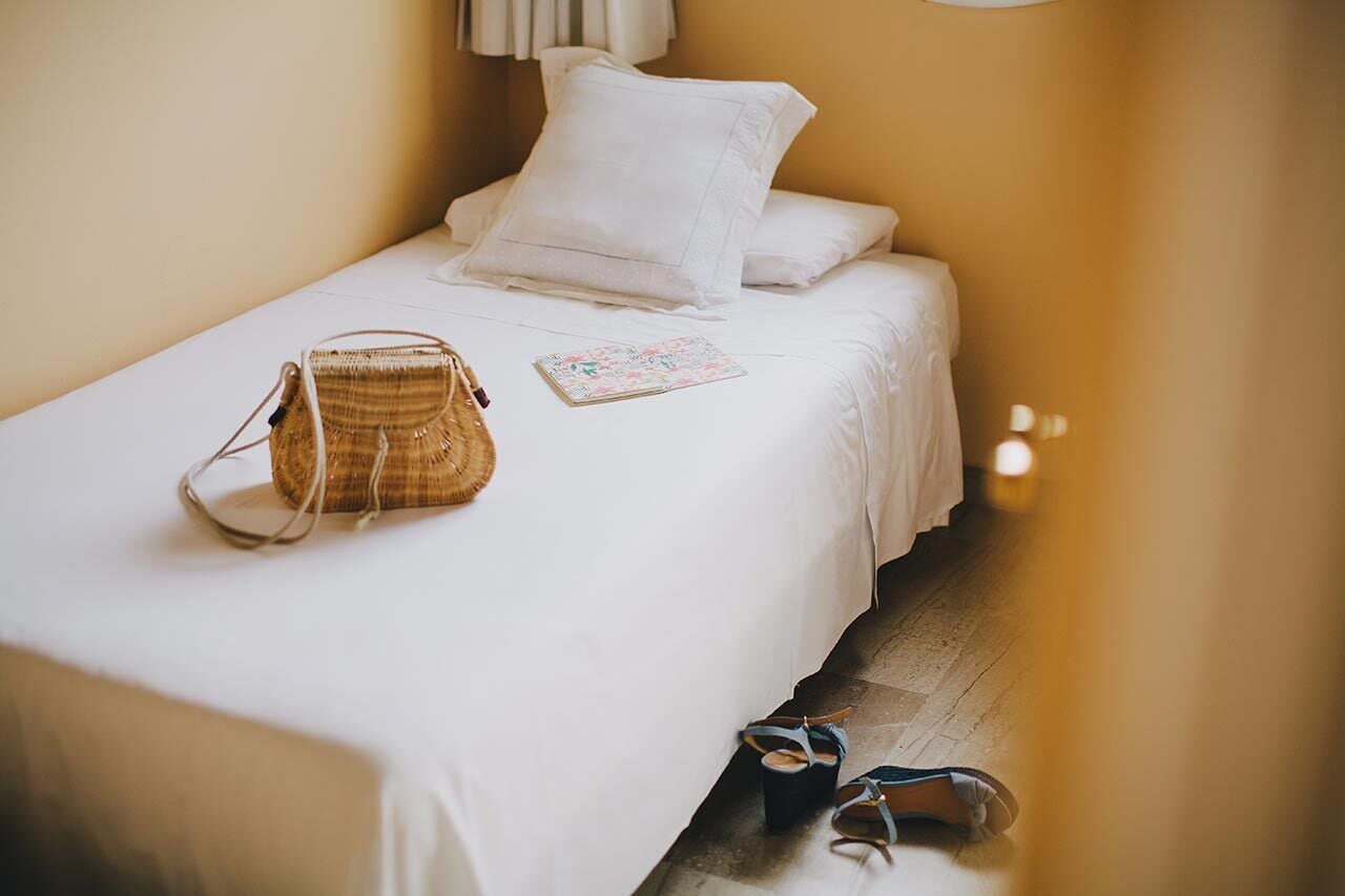 Una cama bien hecha con sábanas blancas, un bolso de mimbre y un par de sandalias en el suelo.