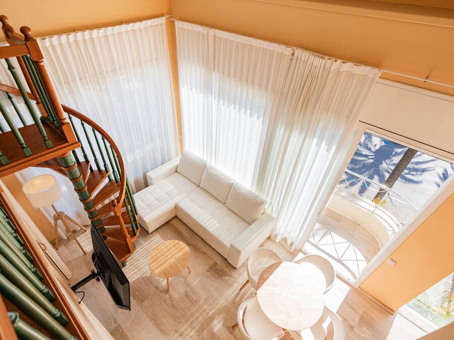 A modern living room with a white sofa, a wooden spiral staircase, a dining area with a round table, and a large window with sheer curtains.