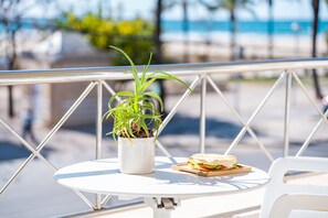 A round white table with a potted plant and a sandwich on it, set against a railing with a view of the beach and ocean.