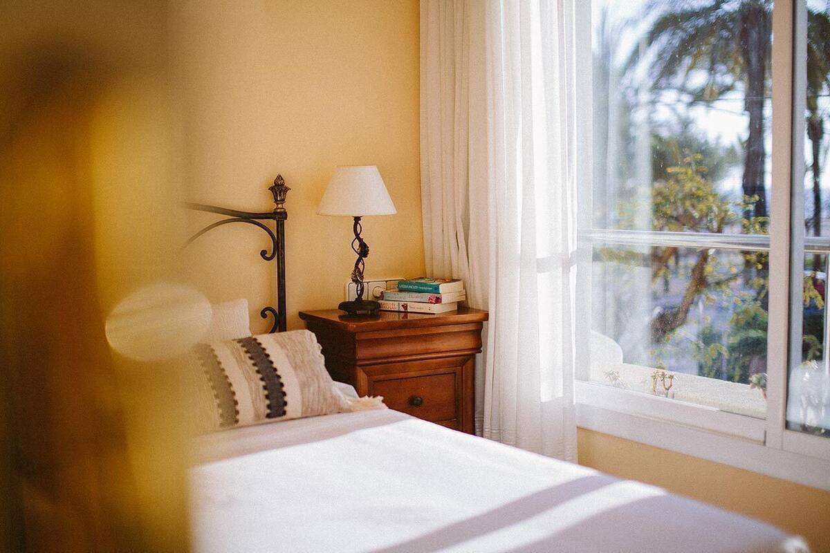 Une chambre à coucher avec un lit, une table de chevet avec une lampe, une fenêtre donnant sur des arbres et un mur agrémenté d’un luminaire décoratif.