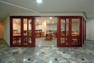 Un salón de conferencias con varias mesas y sillas, suelo de baldosas y puertas de vidrio con marco de madera.