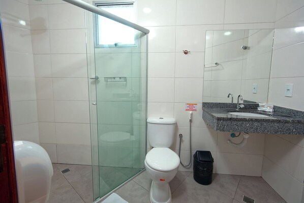 A bathroom with a glass shower enclosure, a toilet, a sink with a mirror above it, and a black trash bin.