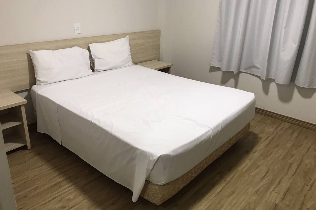 A neatly made bed with white bedding in a room with wooden flooring and a simple headboard.