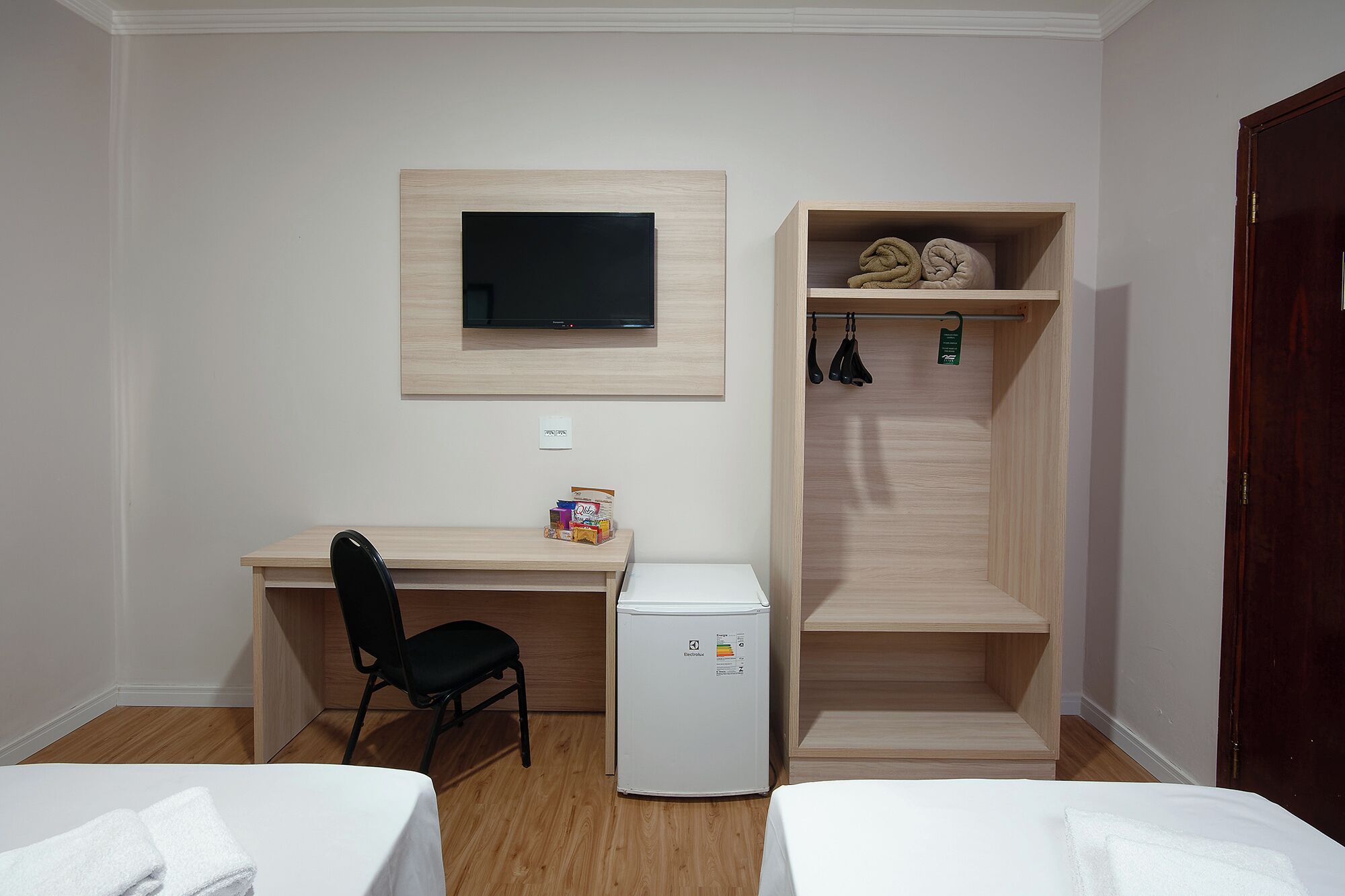 A compact room with a desk, chair, and a small refrigerator. There is a television mounted on the wall above the desk.