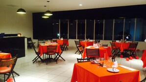 Un restaurante con mesas cubiertas de manteles naranjas, dispuesto en un espacio interior amplio con ventanas grandes y luces de techo.