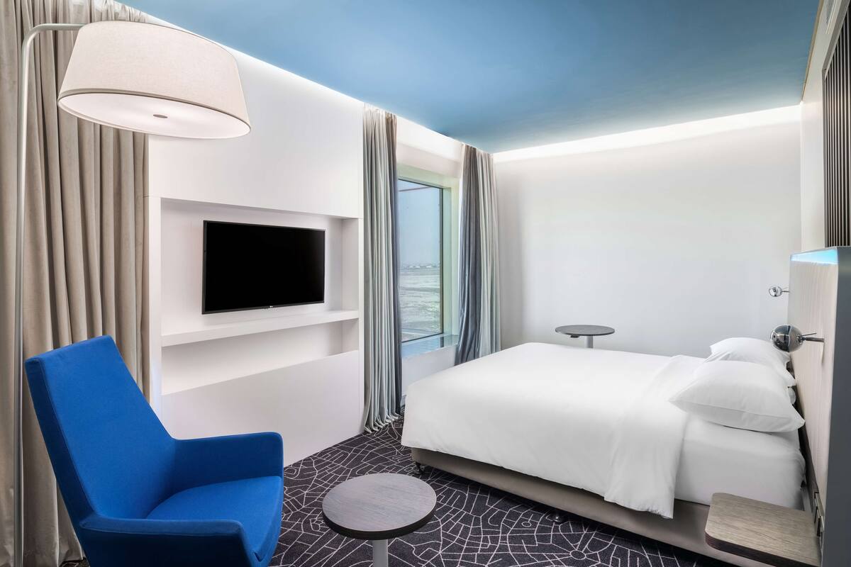 A modern hotel room with a bed, a blue chair, a small table, a TV mounted on the wall, and a window with curtains.