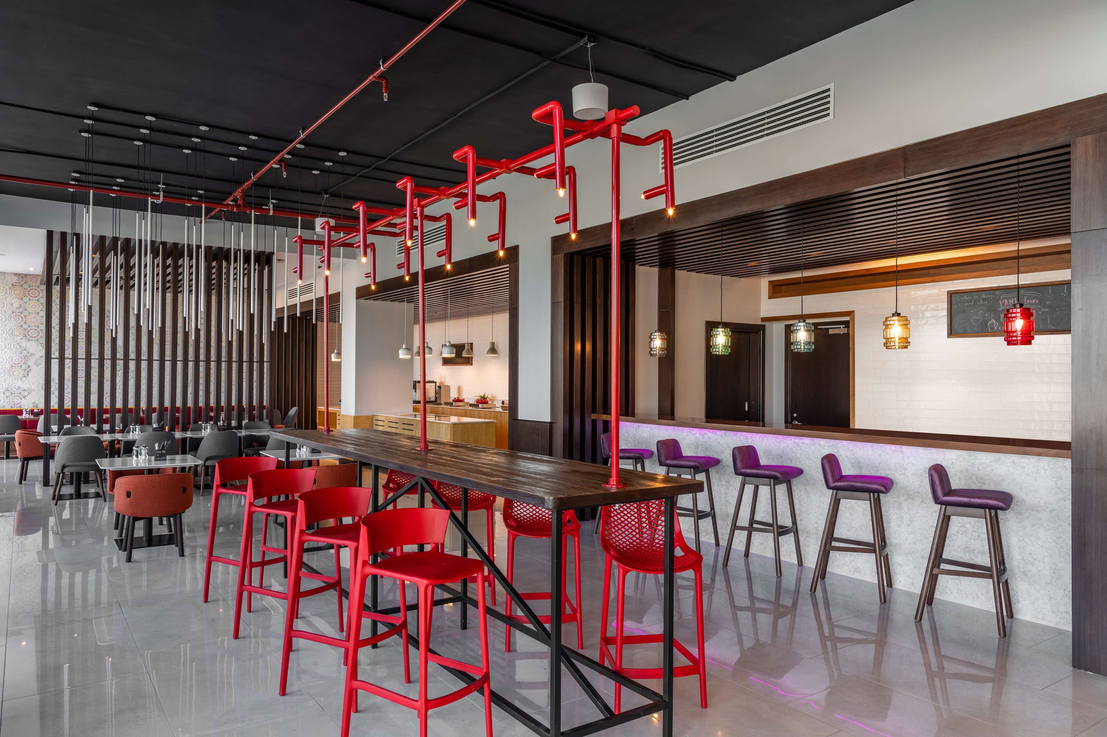 A modern restaurant with a long communal table, red and purple bar stools, and a high ceiling with exposed pipes.