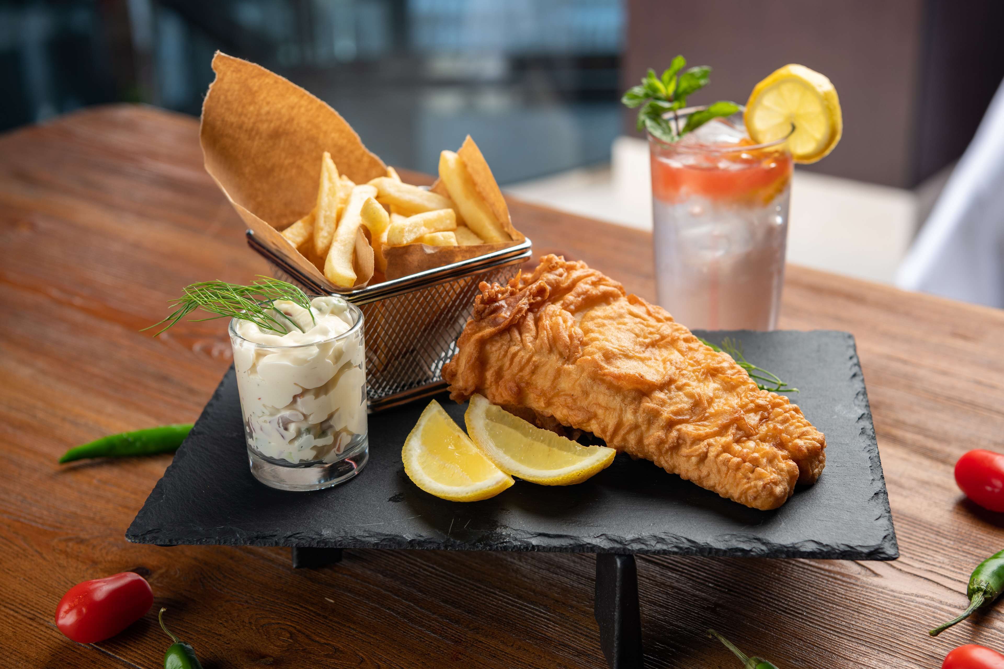 A fish and chips meal on a slate board with lemon wedges and a drink.