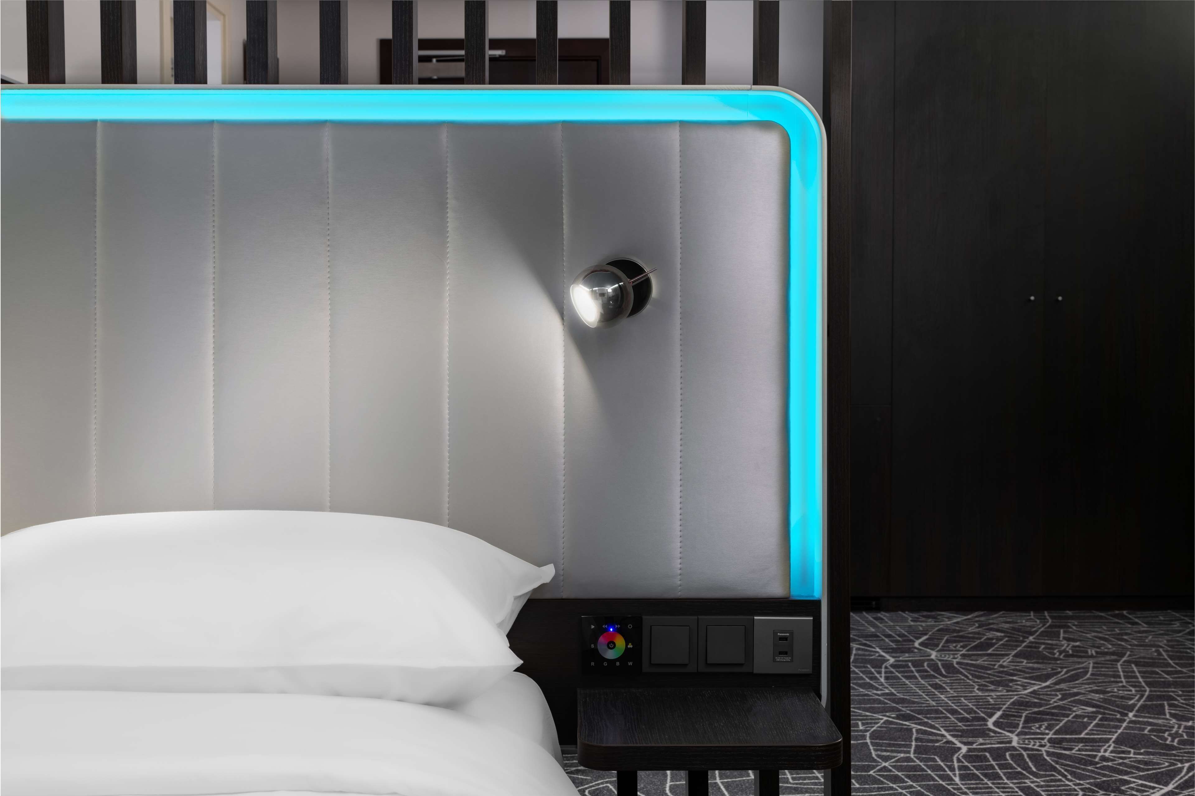 A modern bed with a headboard featuring a light blue accent, a small bedside table with a control panel, and a dark wooden wardrobe in the background.