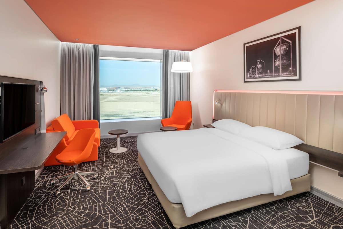 A hotel room with a bed, two orange chairs, a desk, a large window with a view, and a framed picture on the wall.