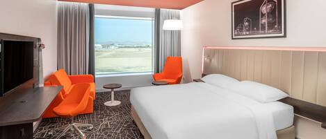 A hotel room with a bed, two orange chairs, a desk, a large window with a view, and a framed picture on the wall.