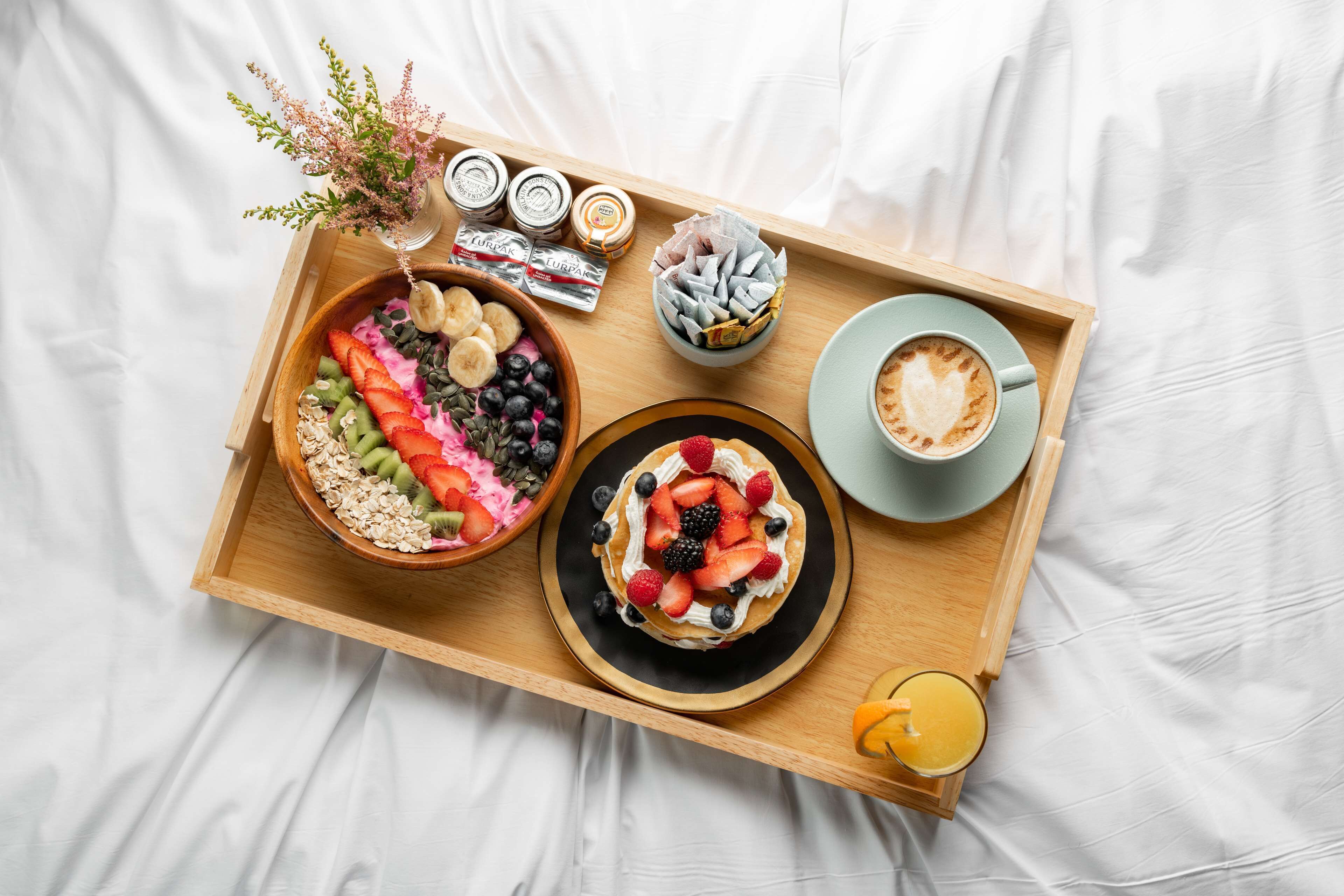 Een houten dienblad met een ontbijt, bestaande uit ontbijtgranen, fruit, een dessert en een kop koffie op een witte laken.