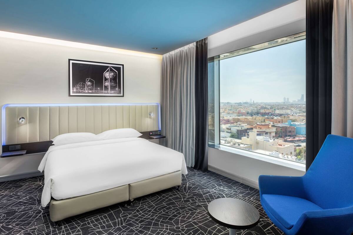 A hotel room with a bed, a chair, a round table, a city view, and a framed picture on the wall.