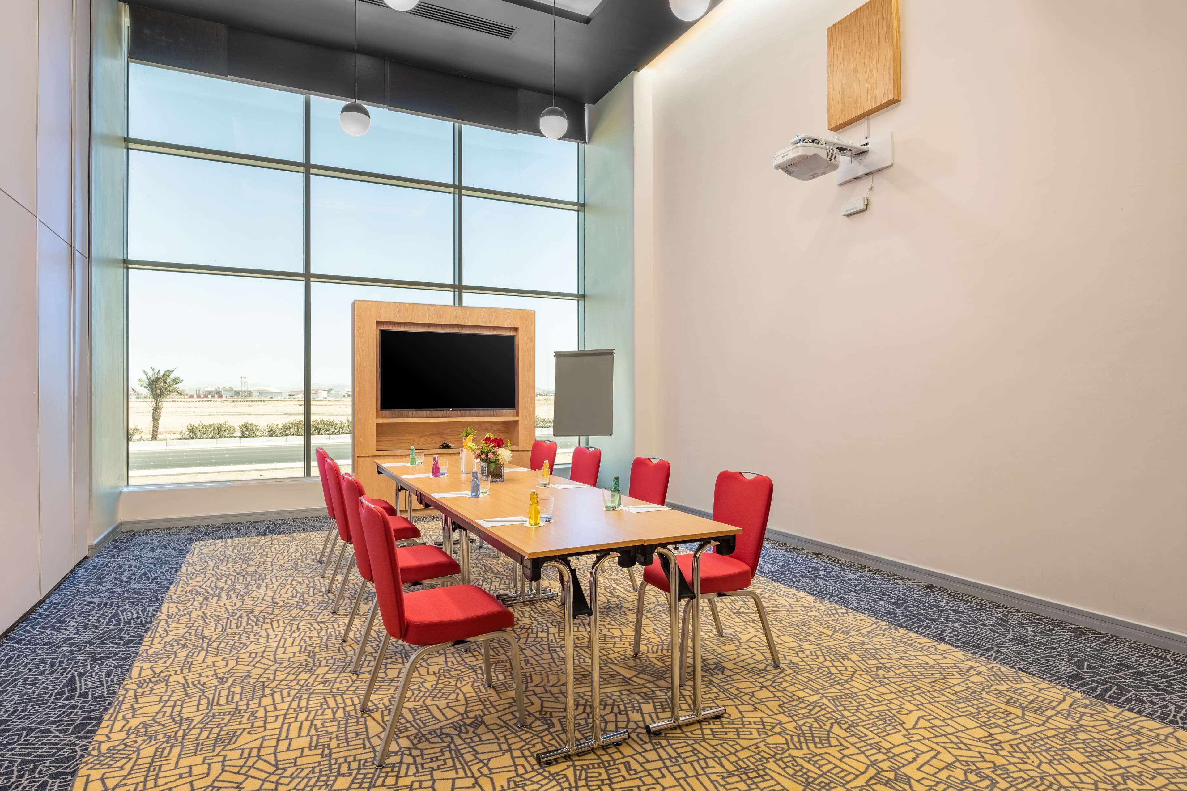 A conference room with a large table, red chairs, a large window, and a flat-screen TV.