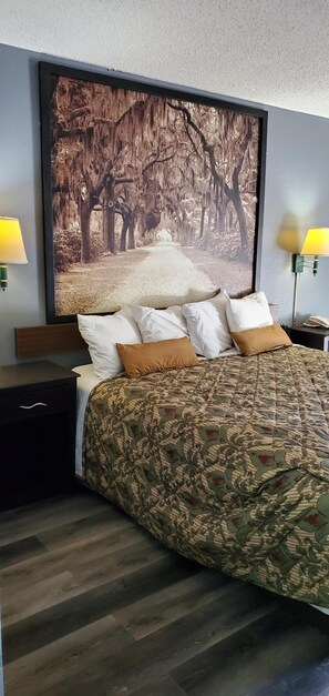 A bedroom with a bed, two bedside tables, a nightstand, a large framed picture of a forest path, and two wall-mounted lamps.