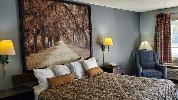 A hotel room with a large bed, two bedside lamps, a chair, a large window with curtains, and a wall-mounted air conditioner. - Palmetto Inn (Estill)