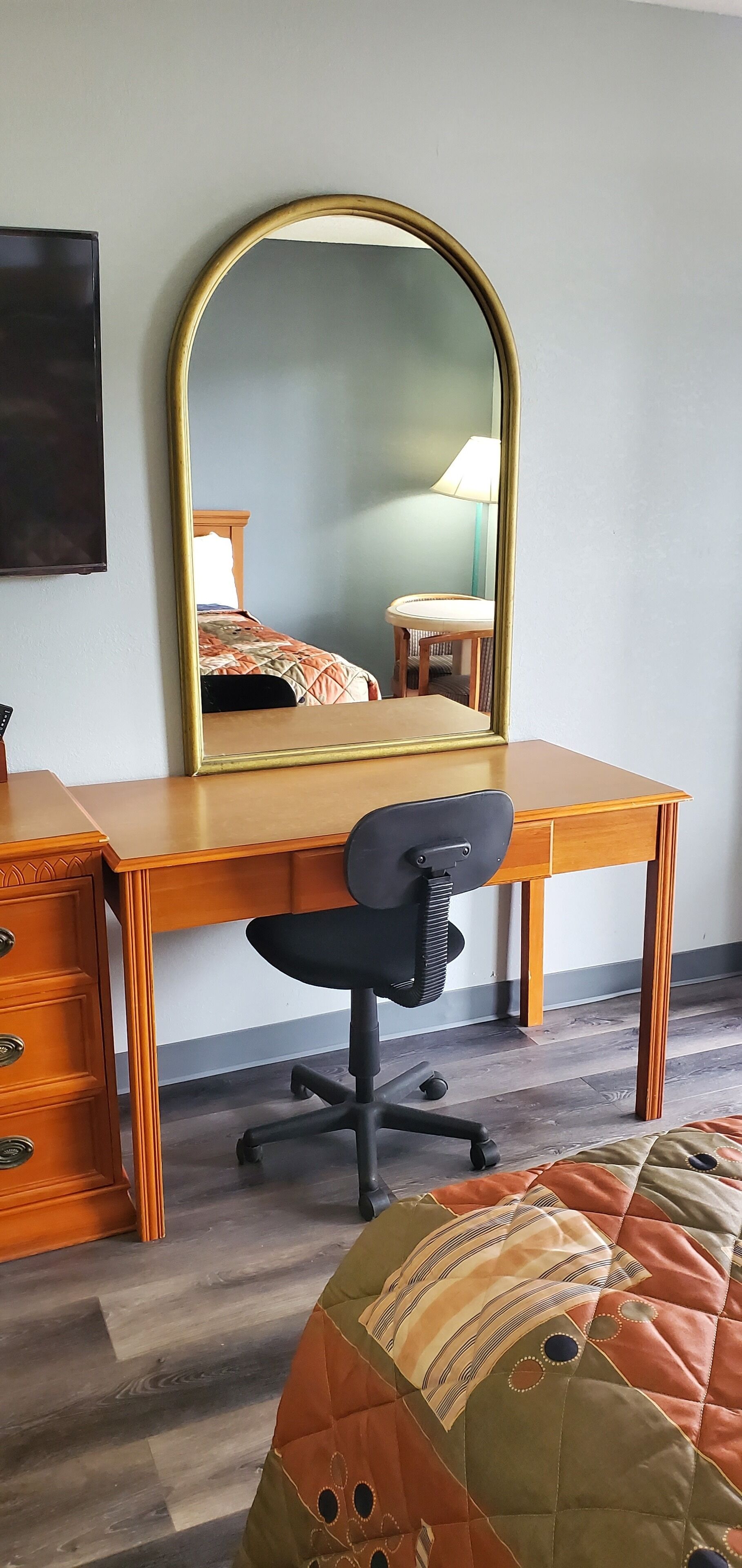 Un dormitorio con un escritorio de madera, una silla de oficina negra, un espejo grande, una cama con colcha y una mesita de noche con lámpara.