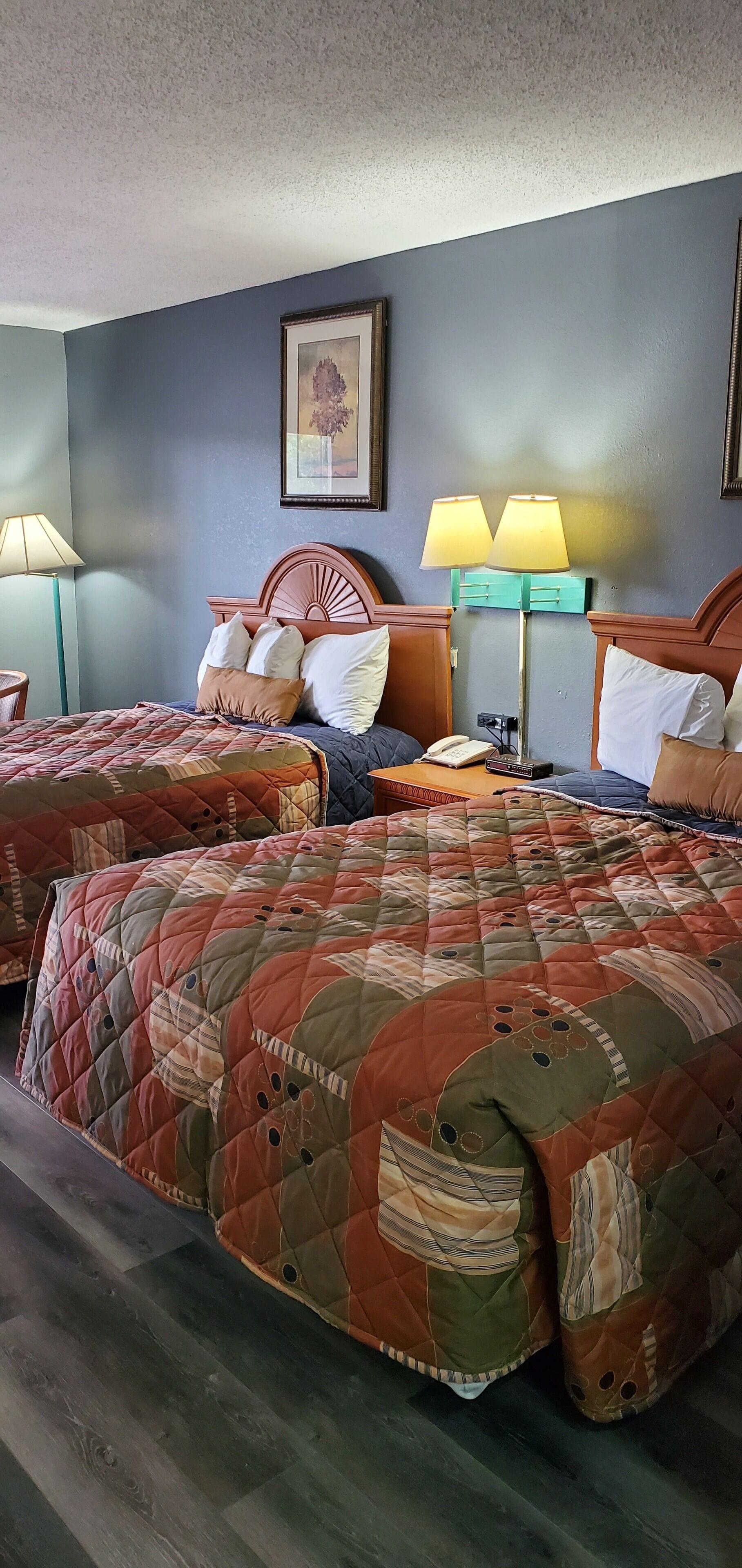A hotel room with two beds, each with a quilted bedspread, a wooden headboard, and a bedside table with a lamp.