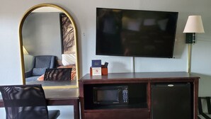 A hotel room with a flat-screen TV mounted on the wall, a desk with a microwave, a chair, and a lamp. - Palmetto Inn (Estill)
