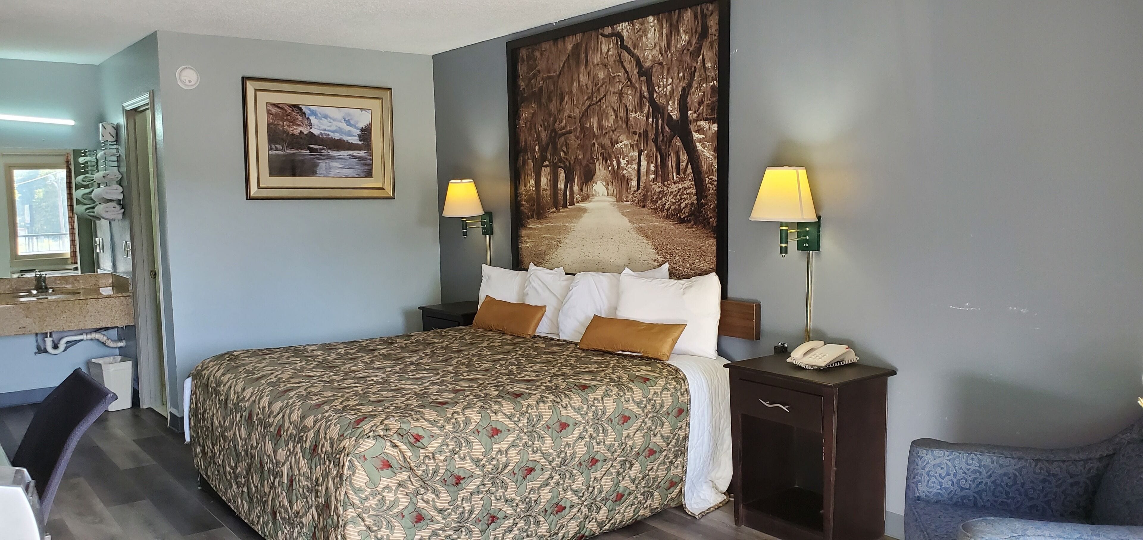 A hotel room with a large bed, two bedside lamps, a nightstand with a phone, a framed picture on the wall, and a window with a view of trees.