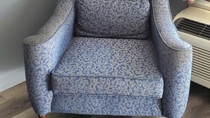 A blue armchair with a patterned fabric, positioned against a grey wall with a white lamp and plaid curtains in the background. - Palmetto Inn (Estill)