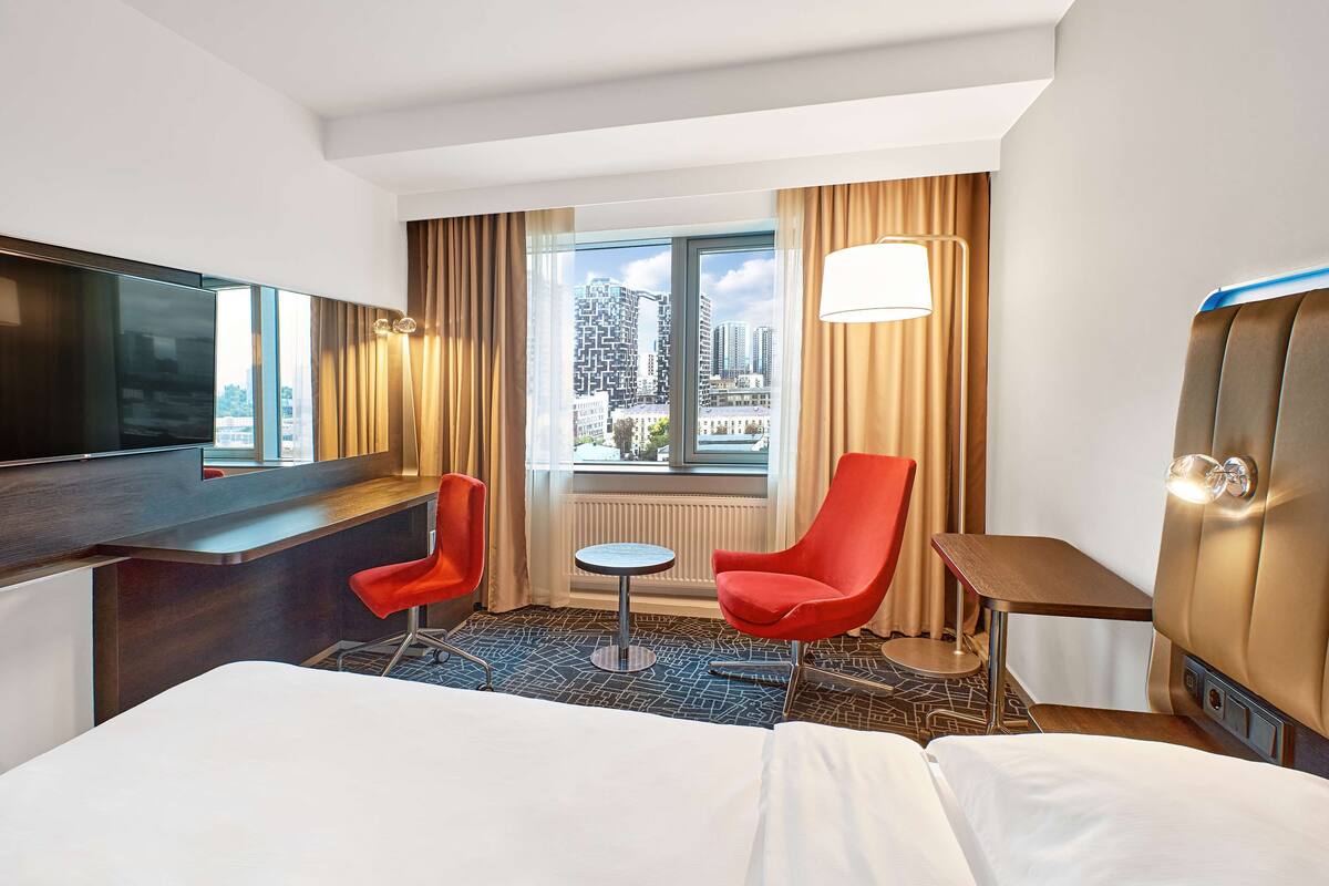 Ein Hotelzimmer mit Bett, Fernseher, kleinem Tisch, zwei roten Stühlen, einer Lampe und Blick auf Gebäude durch das Fenster.