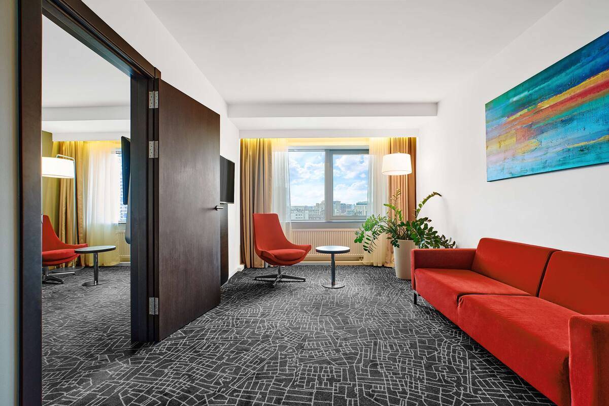 Ein Hotelzimmer mit rotem Sofa, rotem Sessel, einem kleinen Tisch, einem großen Fenster mit Vorhängen, Blick auf die Stadt und einem farbenfrohen, abstrakten Gemälde an der Wand.