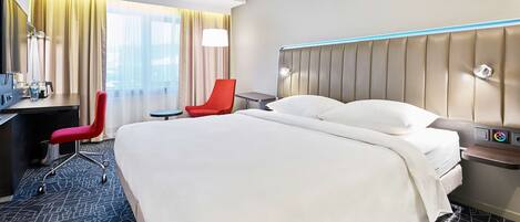 A hotel room with a large bed, a desk with a chair, a red chair, a bedside table with a lamp, and a large window with curtains.
