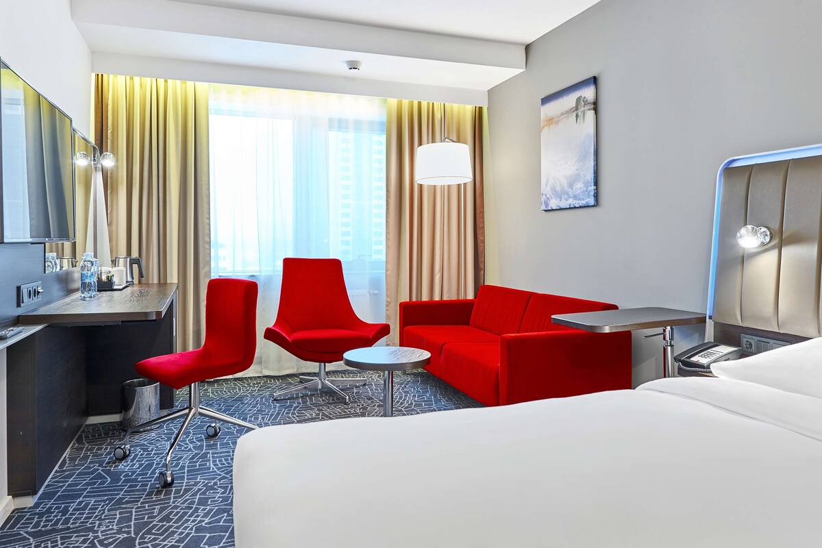 Ein Hotelzimmer mit einem Bett, zwei roten Stühlen, einem Schreibtisch mit Computer und einem kleinen Tisch.