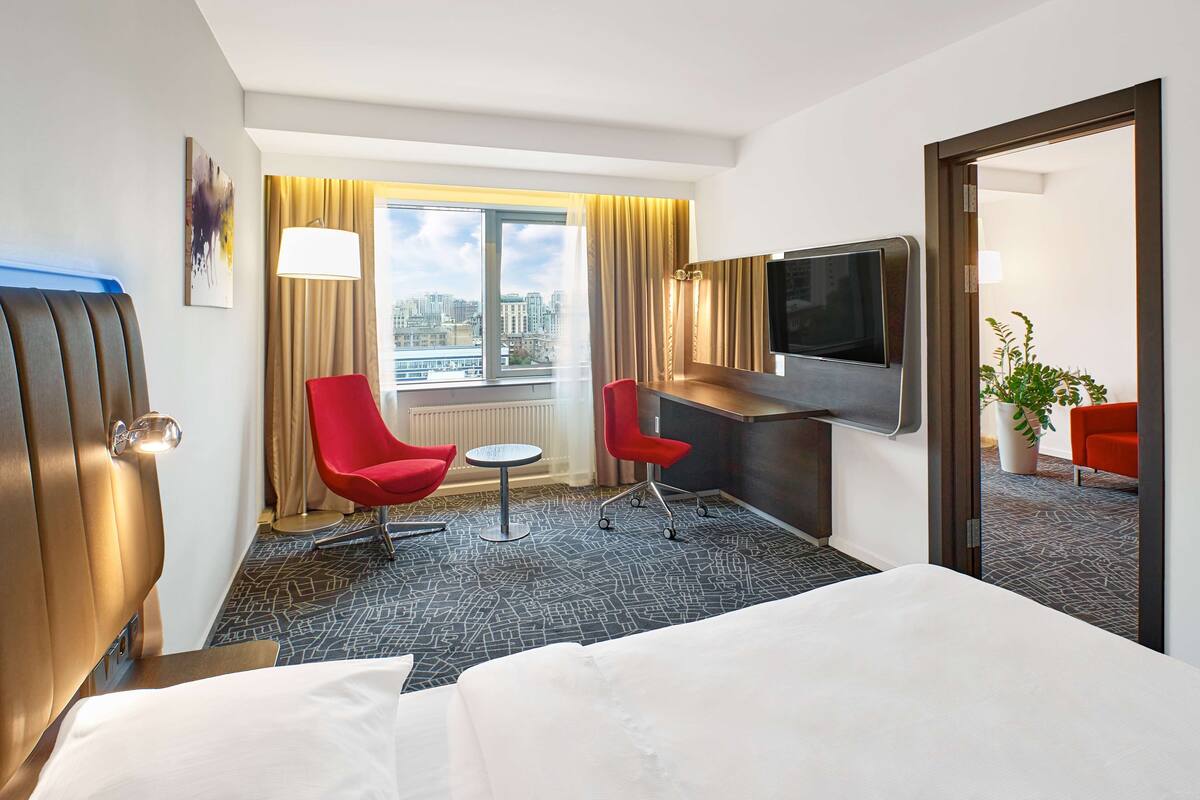 Ein Hotelzimmer mit einem Bett, einem Schreibtisch, zwei roten Sesseln, einem Fernseher, einem Fenster mit Blick auf die Stadt und einer Zimmerpflanze.
