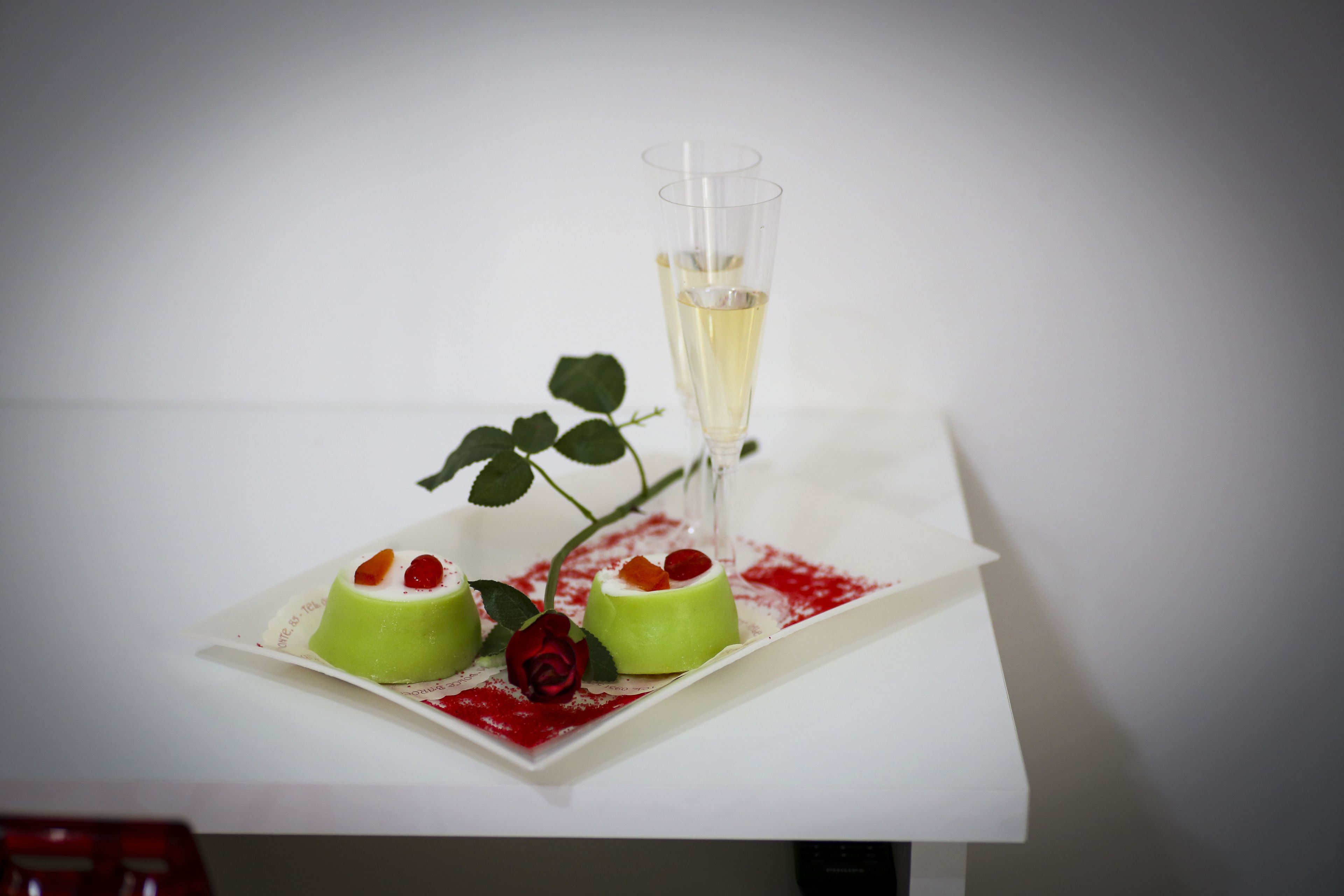 Un plato de postre con dos bombones verdes cilíndricos, una rosa y una copa de champán sobre una mesa blanca.