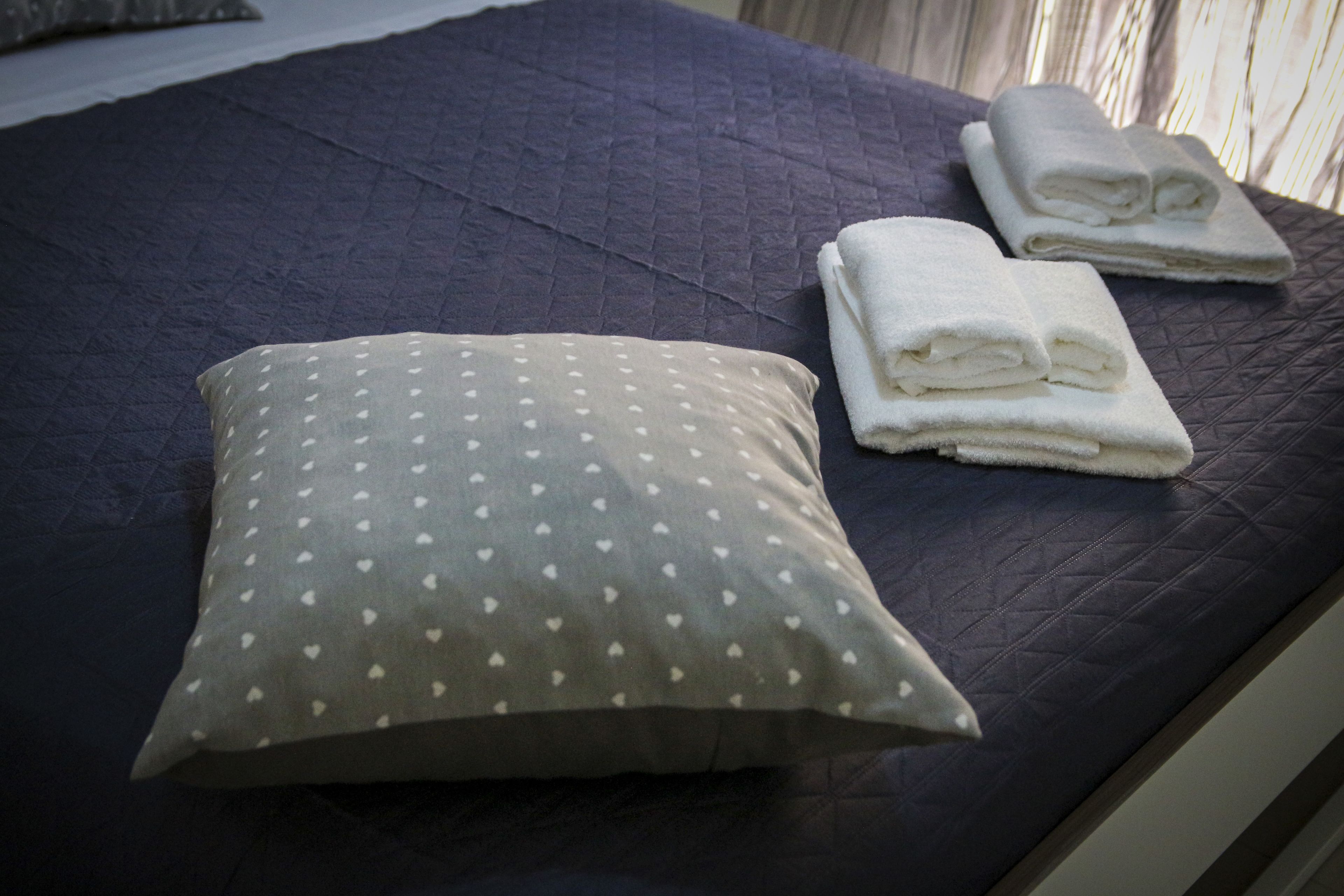 A bed with a dark quilted cover, a pillow with a heart pattern, and two folded towels.