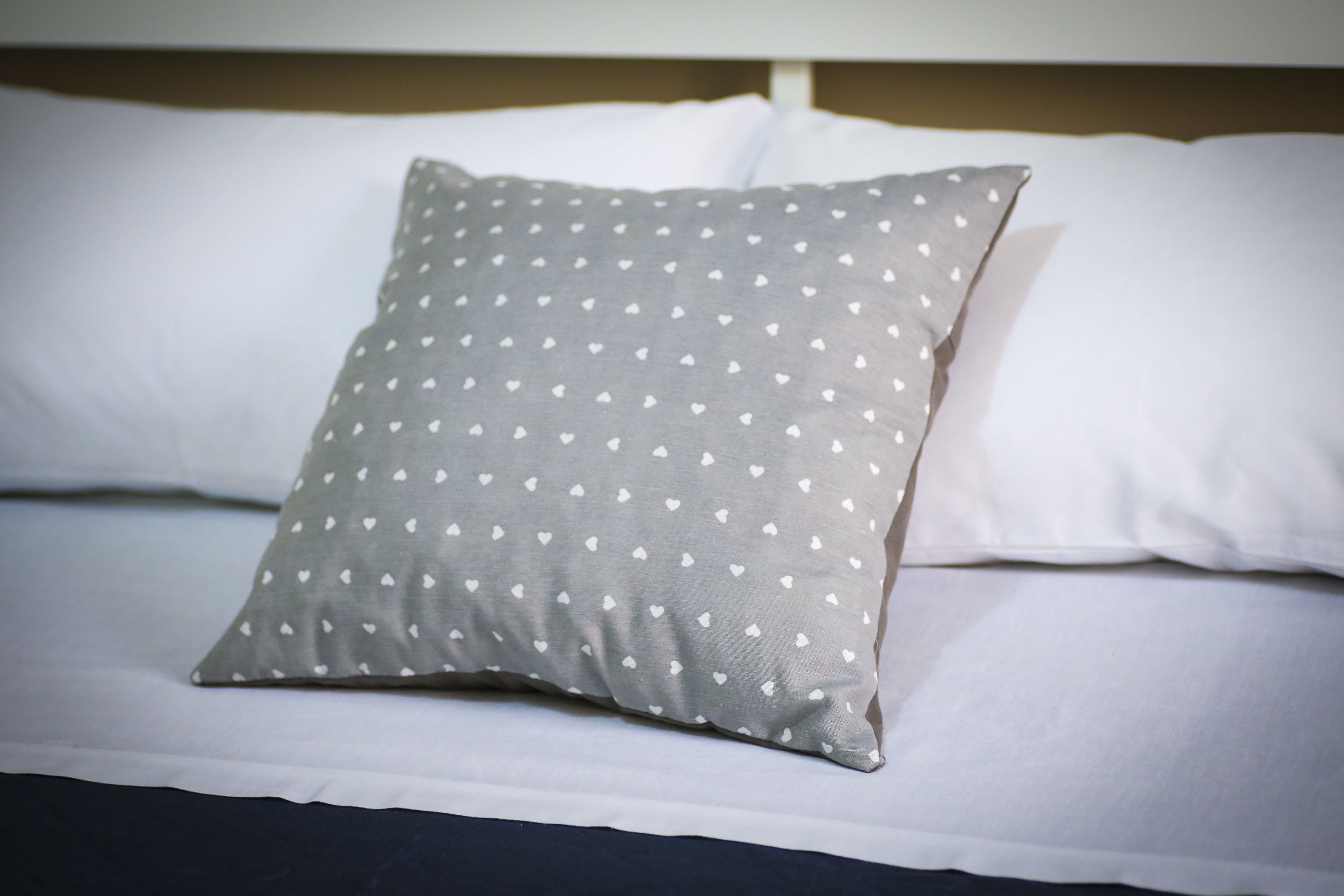 A bed with white bedding and a gray pillow with heart patterns.