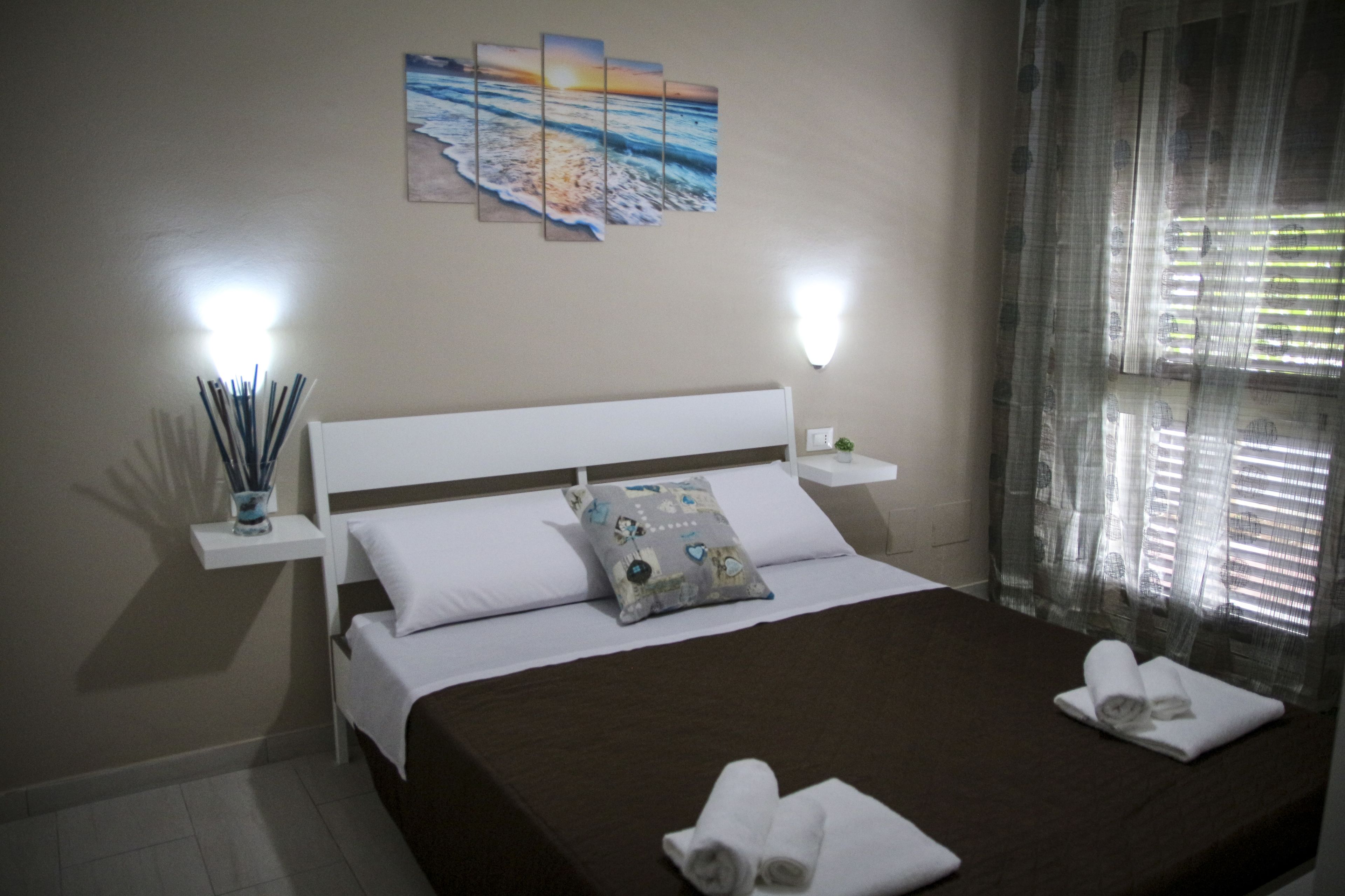 A neatly made bed with white and brown linens, a decorative pillow, and rolled towels on the bed. There are two wall-mounted lights and a painting of a beach scene above the bed.