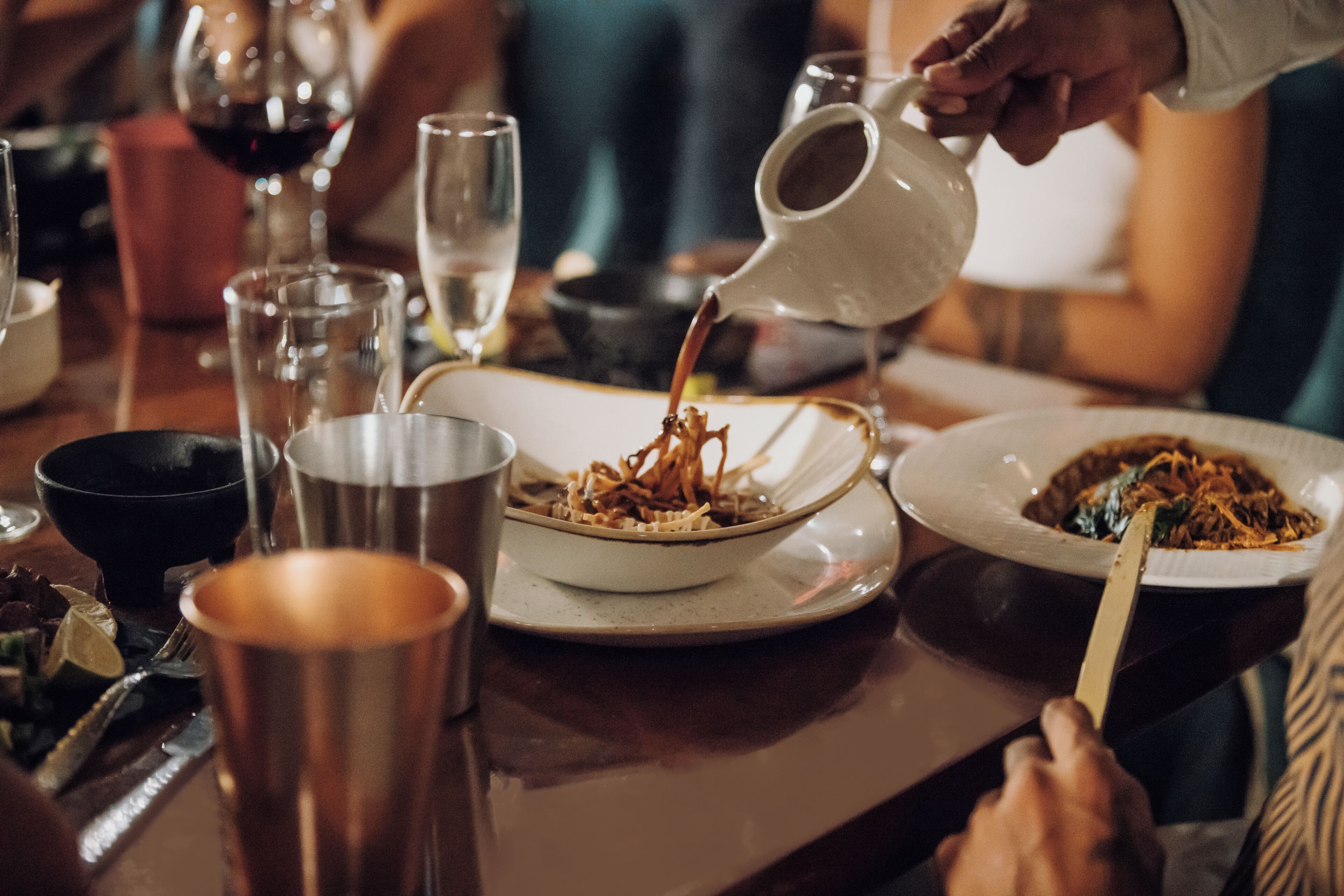 A dining table with various dishes, a pitcher pouring liquid, and a person using chopsticks.