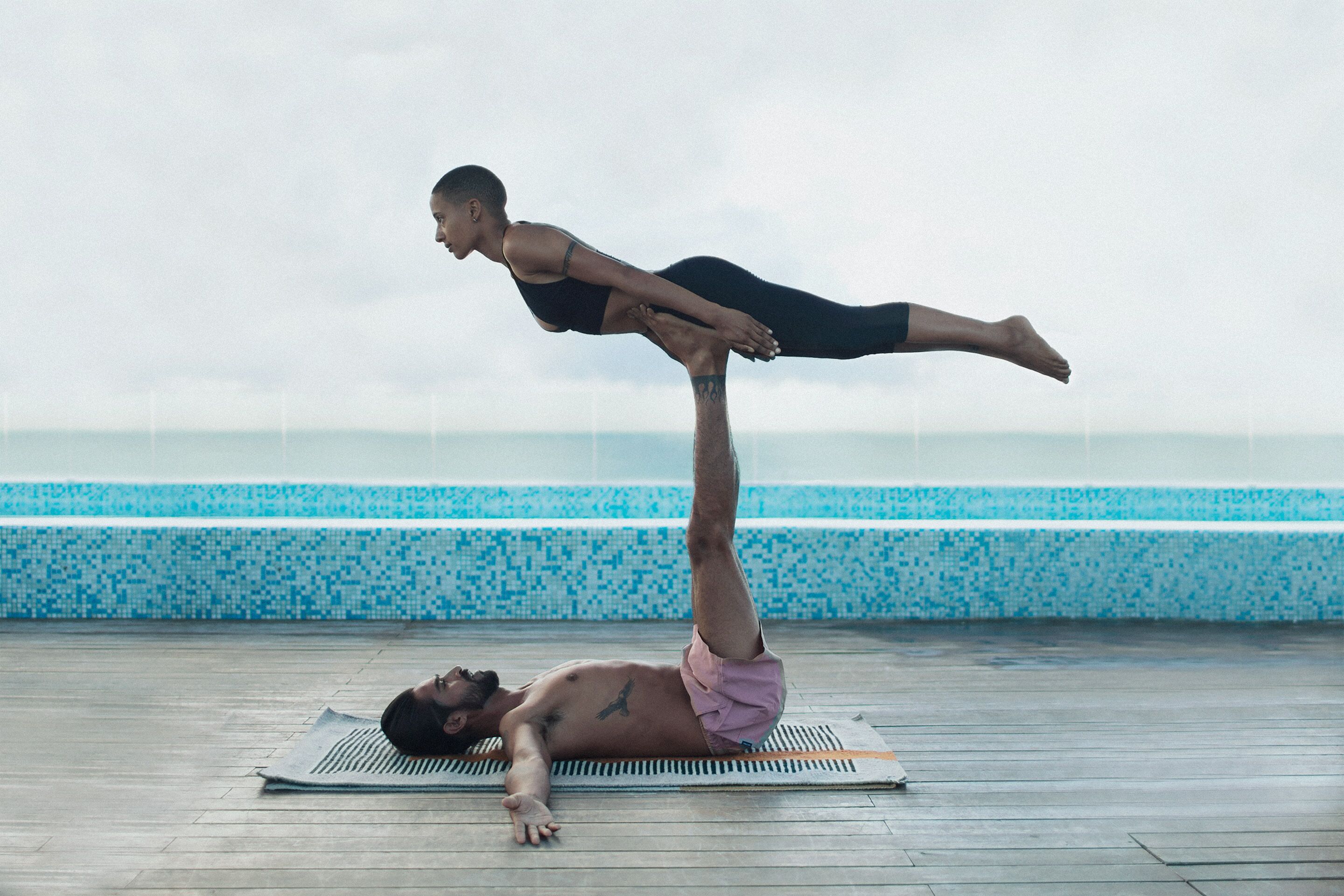 A person performing a yoga pose on a mat by a poolside with a tiled wall and a view of the water.