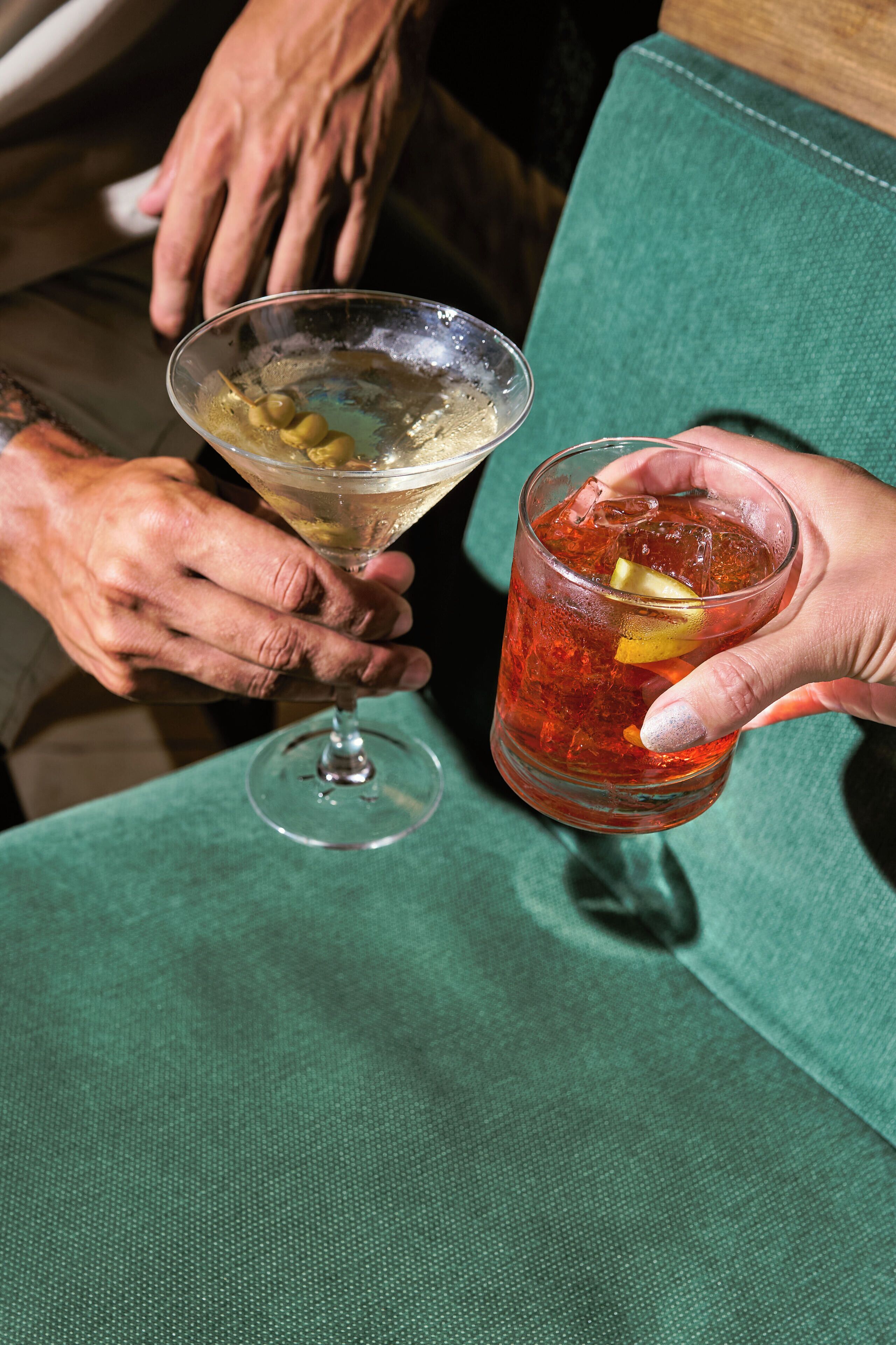 Deux personnes tenant des verres, l’une un verre à martini et l’autre un verre à whisky, avec une chaise verte à l’arrière-plan.
