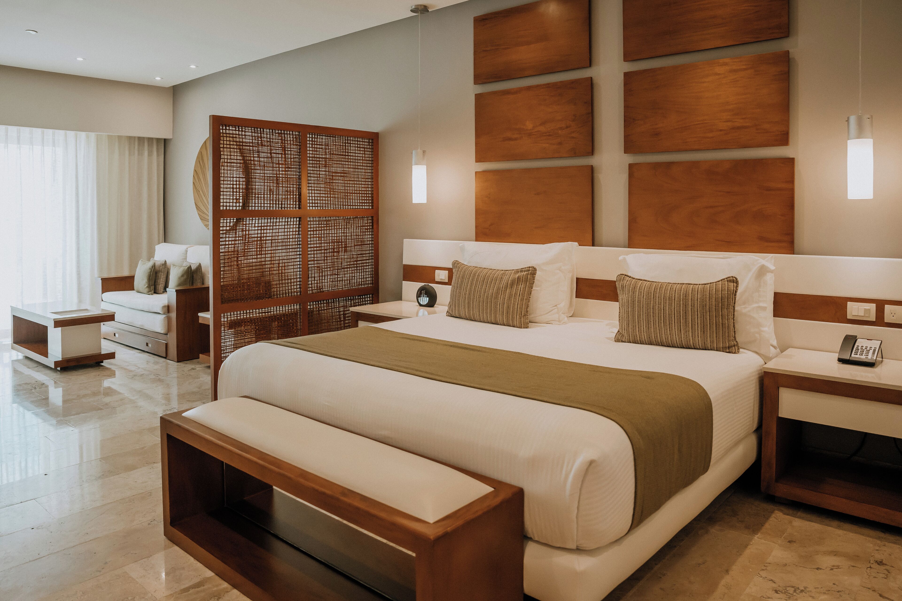 Una habitación de hotel moderna con una cama grande, cabecera de madera, mesitas de noche, una partición tejida y un ventanal con cortinas.