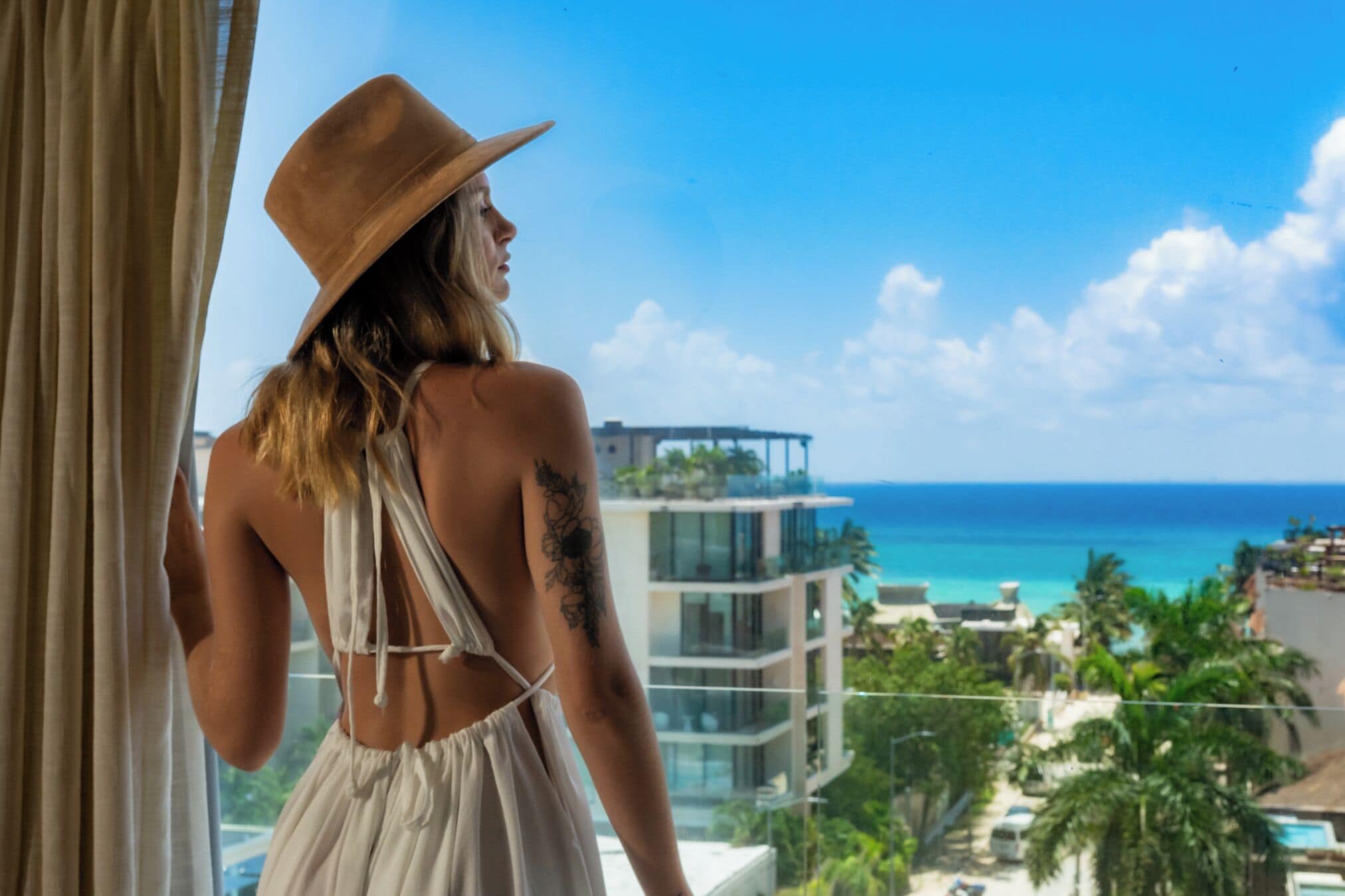 A person in a white dress and hat looking out at a coastal view with buildings and palm trees.