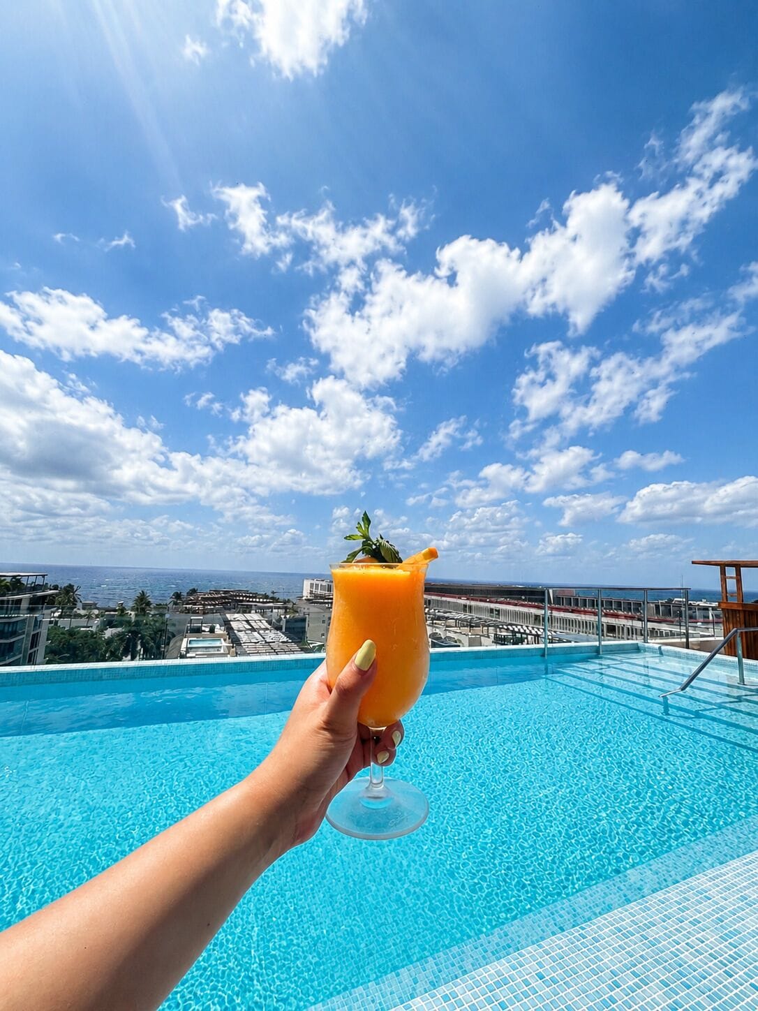 Una mano sosteniendo un vaso de jugo de naranja sobre una piscina, con un cielo azul despejado y edificios a lo lejos.