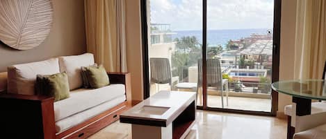 A modern living room with a sofa, a glass coffee table, and a view of a resort with a pool and buildings in the distance.