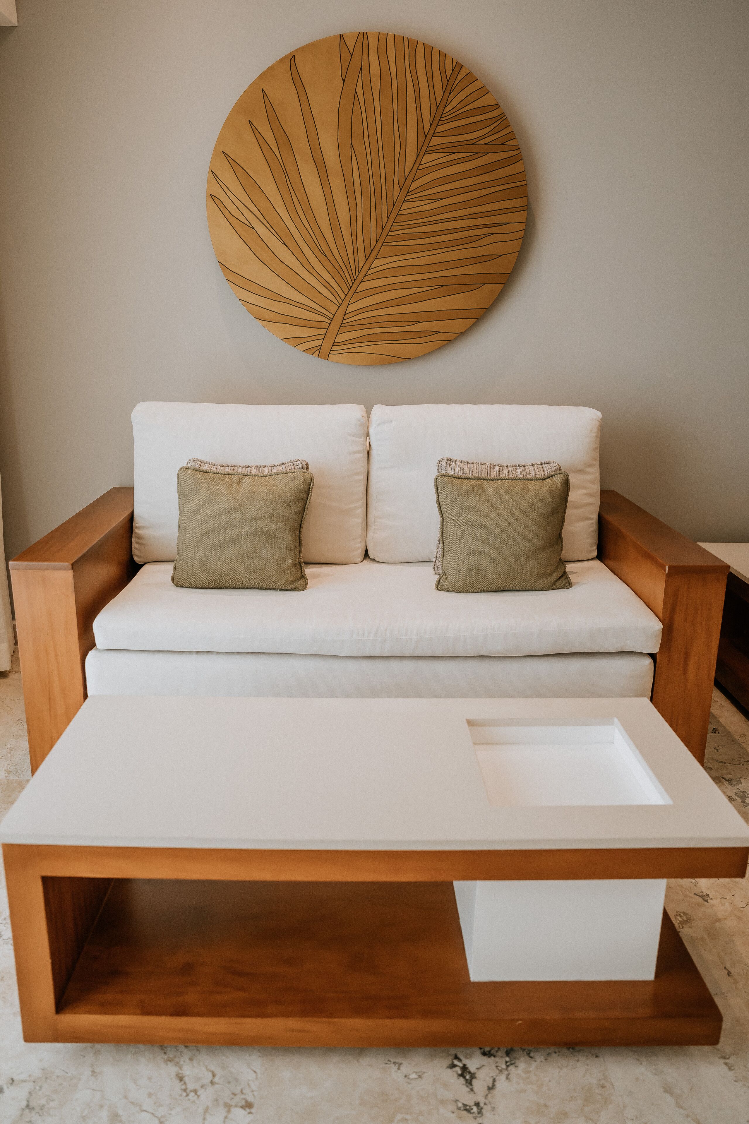 Ein modernes Wohnzimmer mit einem weißen Sofa, zwei grünen Kissen, einem Holztisch und einem großen Wandbild mit Blättern.