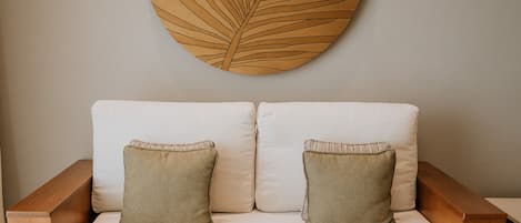 Ein modernes Wohnzimmer mit einem weißen Sofa, zwei grünen Kissen, einem Holztisch und einem großen Wandbild mit Blättern.