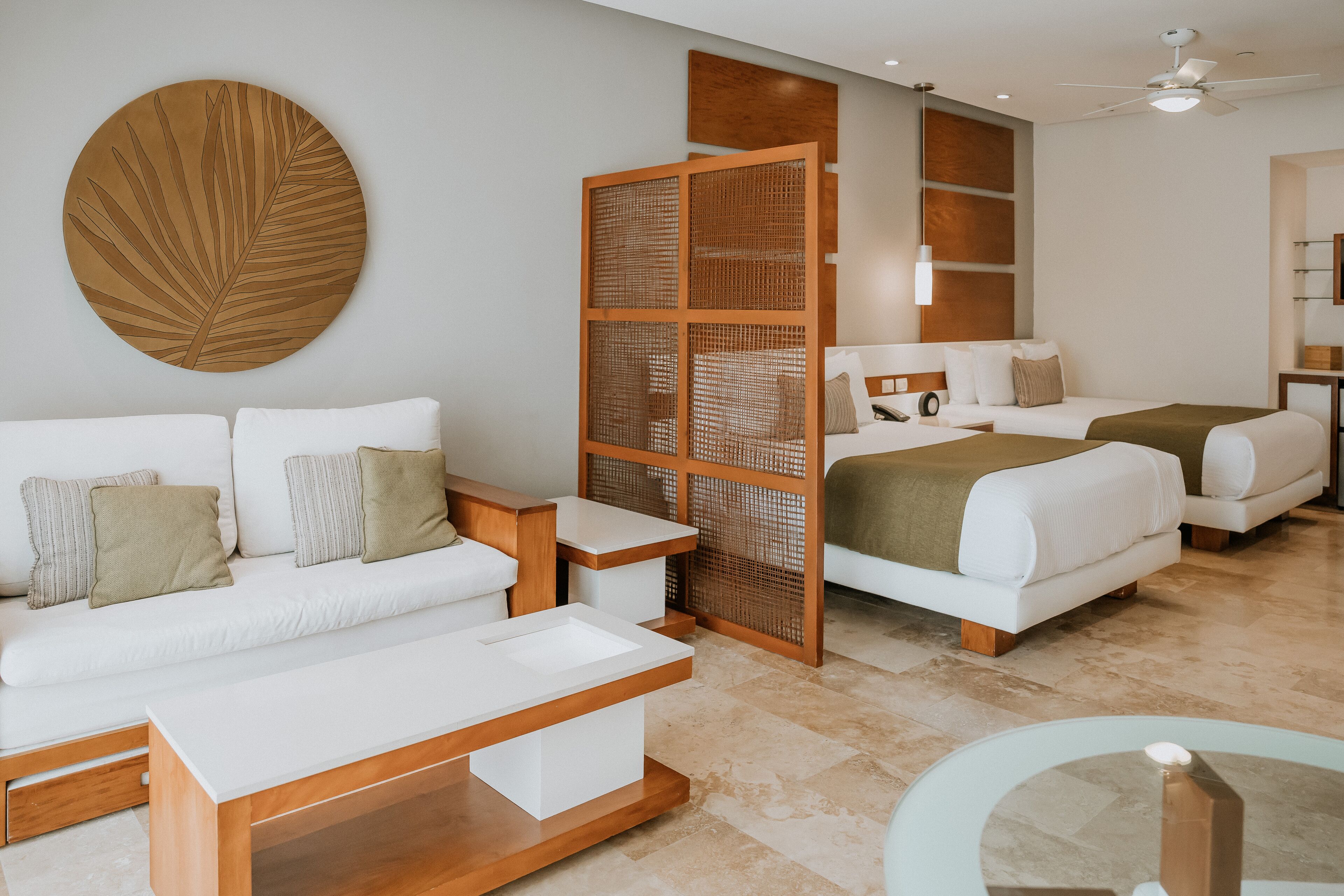 A modern hotel room with a white sofa, a wooden coffee table, a bed with a green blanket, and a large decorative wall piece.