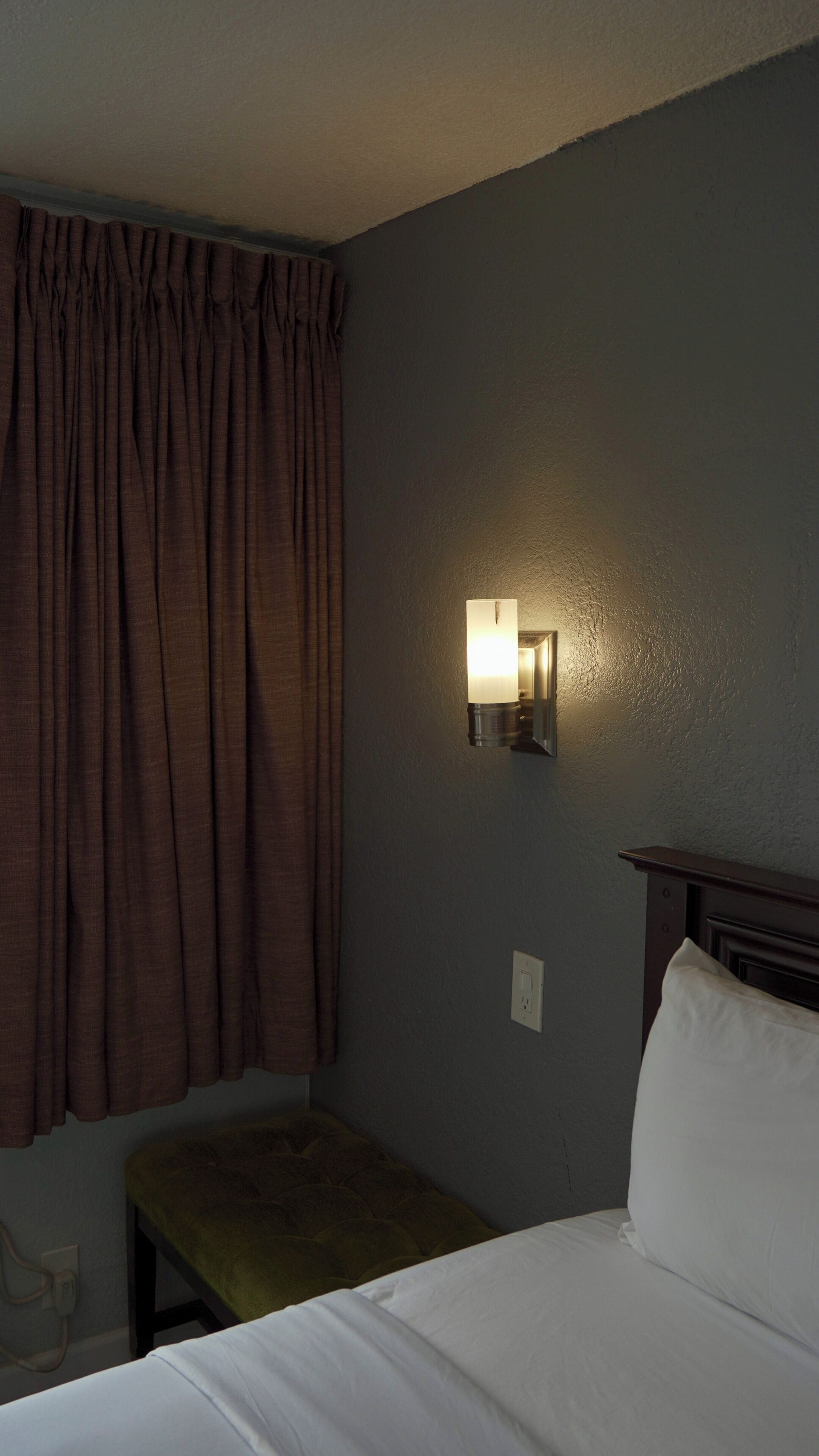 Habitación de hotel con una cama, una mesita de noche, una lámpara de pared y un ventanal con cortinas.