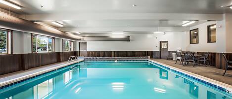 Un'ampia piscina interna con bordo piastrellato, un'area lounge con tavoli e sedie, e vista sull'esterno tramite ampie finestre.