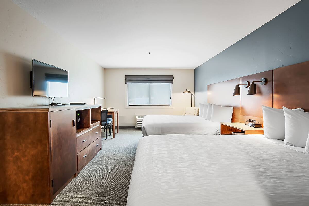 A hotel room with two beds, a desk, a TV, and a window with blinds.
