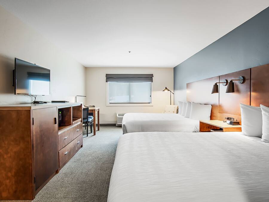 A hotel room with two beds, a desk, a TV, and a window with blinds.