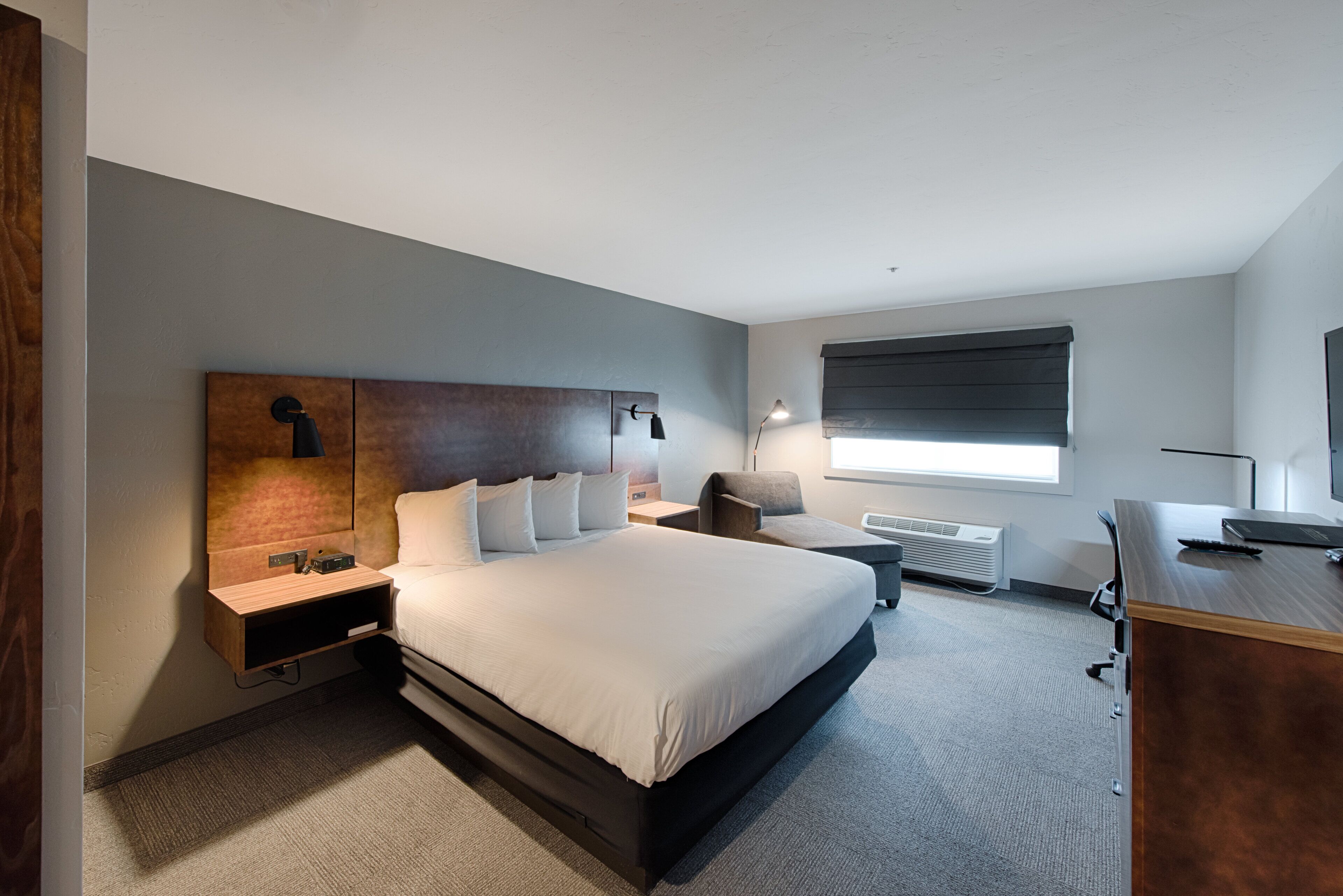 Una habitación de hotel moderna con una cama grande, un escritorio, una silla, una mesita de noche y una ventana con persianas.