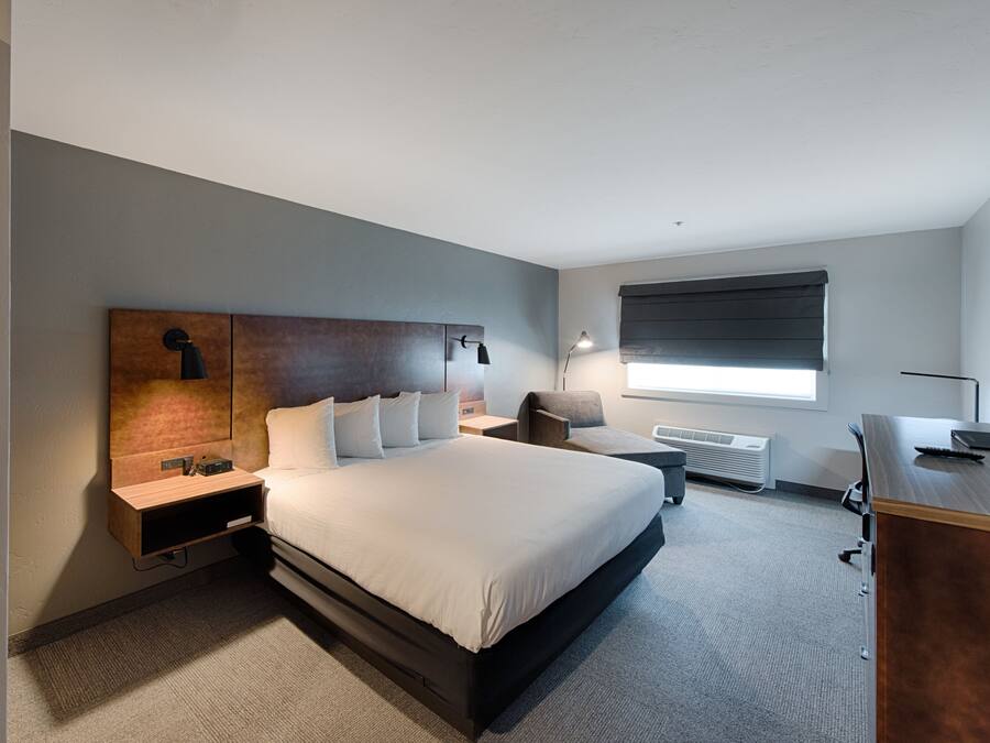Una habitación de hotel moderna con una cama grande, un escritorio, una silla, una mesita de noche y una ventana con persianas.
